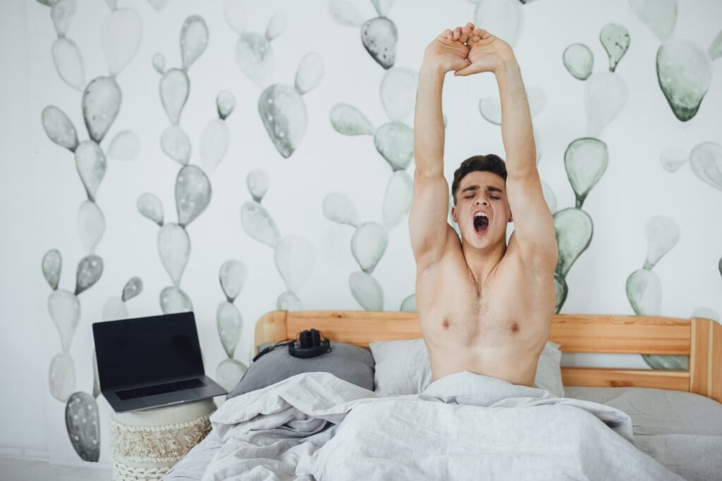 ఉదయాన్నే మంచం మీద లేచి ఒళ్ళు వంచుతున్న యువకుడు – ఆరోగ్యకరమైన జీవనశైలి సూచన